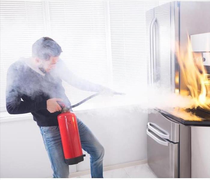 Young Man Using Red Fire Extinguisher To Stop Fire Coming From Oven In Kitchen