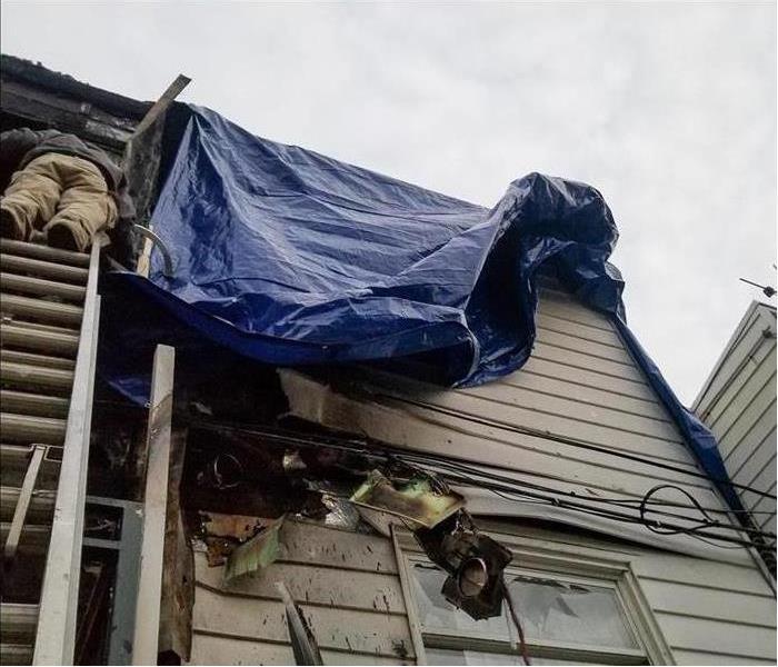 A person on a ladder outside a house that has been exposed to fire damage, blue tarp on roof.
